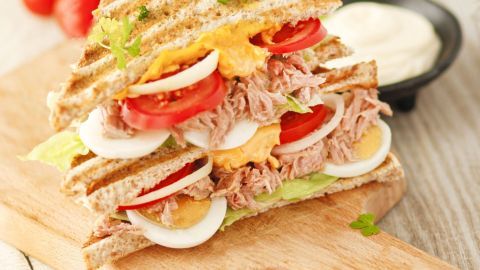 Ein großzügig belegtes Thunfisch-Sandwich aus Vollkornbrot liegt auf einem Holzbrett, umgeben von Kirschtomaten und einer Schüssel Mayonnaise.