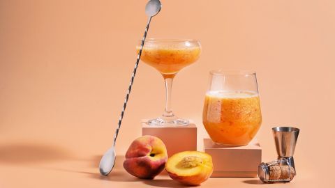 Zwei Gläser mit Pfirsich-Bowle stehen auf einer elegant gestalteten Oberfläche, umgeben von frischen Pfirsichen und einem Cocktail-Shaker.