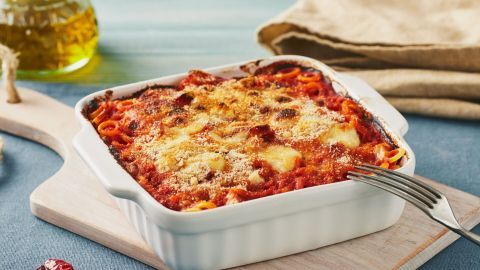 Eine weiße Auflaufform enthält eine überbackene Pasta mit goldbrauner Kruste, umgeben von einem Holzbrett und Ölflasche.