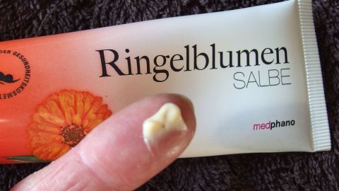 Eine Tube Ringelblumencreme wird mit einem Finger gehalten, bereit zur Anwendung bei einer roten, entzündeten Nase.