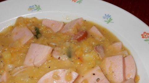 Herzhafte Linsensuppe serviert in einer weißen Schüssel mit Stückchen von Wurst und Gemüse, auf einem Holztisch.