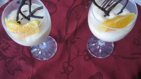 Zwei Weingläser mit Orangencreme, dekoriert mit Orangenscheiben und Schokoladengitter, stehen auf einem roten Tischtuch.