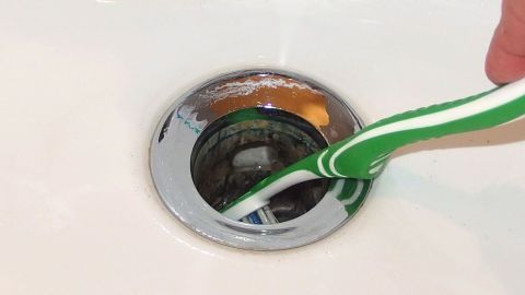 Eine Zahnbürste mit grün-weißen Borsten wird in einen Abfluss gesteckt, in einem hellen Waschbecken sichtbar.