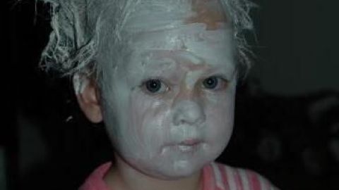 Ein Kind mit Penaten-Creme im Haar und Gesicht trägt einen gestreiften Pyjama, sitzt in einem dunklen Raum.