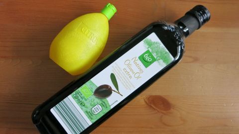 Eine Flasche Olivenöl liegt neben einer gelben Zitrone auf einer holzfarbenen Oberfläche. Beide Zutaten sind für kaschmirweiches Haar.