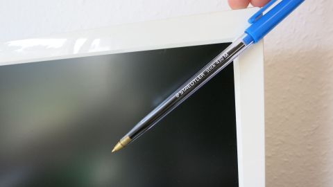 Ein blauer Kugelschreiber wird von einer Hand an den Rand eines schwarzen TFT-Monitors gehalten.