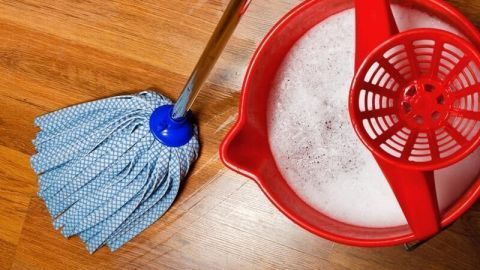 Ein Mopp steht neben einem roten Eimer mit Seifenwasser auf einem Laminatboden, bereit zum Putzen.