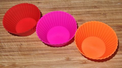 Backformen aus Silikon kann man zur Aufbewahrung richtig klein machen und so ziemlich viel Platz sparen. Außerdem klebt nichts fest. Silikon-Backformen in Rot, Pink und Orange stehen auf einer Holzoberfläche, bereit zum Backen von kleinen Leckereien.