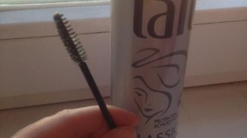 Eine Hand hält eine Wimpernbürste neben einer Dose Haarspray, die für das Fixieren von Augenbrauen verwendet wird.