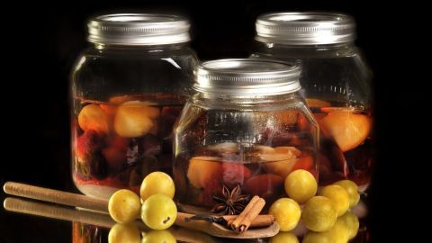 Einmachgläser gefüllt mit Rumtopf-Früchten stehen auf glänzendem Untergrund, umgeben von gelben Mirabellen und Zimtstangen.