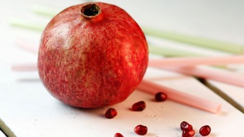 Ein leuchtend roter Granatapfel liegt auf einem weißen Tisch, umgeben von pinken Stäbchen und einigen herausgefallenen Kernen.