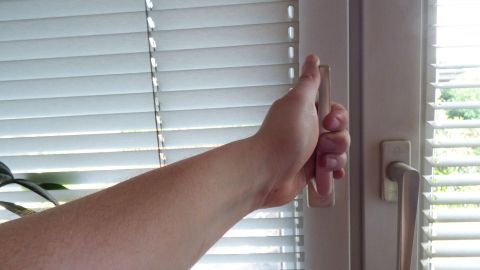 Eine Hand öffnet ein Fenster mit geschlossenen Jalousien, um frische Luft in die Wohnung zu lassen, während es windstill ist.