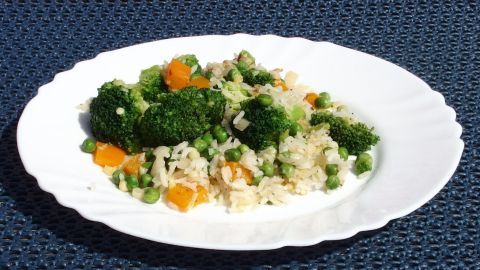 Eine gefüllte weiße Platte zeigt eine bunte, vegane Paella mit Brokkoli, Erbsen und Karotten, serviert auf einem blauen Tisch.