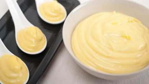 Kremiger Vanillepudding in einer Schüssel, umgeben von Löffeln mit ähnlichem Pudding auf einem schwarzen Teller.