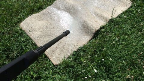Ein Teppich liegt auf dem Gras, während ein Hochdruckreiniger ihn von Schmutz befreit und auffrischt.