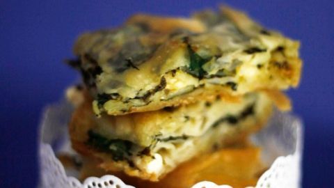 Schichten von Spinat-Pita liegen auf einem eleganten weißen Tortenteller, umgeben von einem blauen Hintergrund.