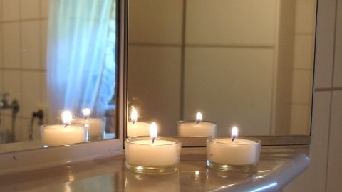 Spiegel beschlagen beim Duschen nicht, wenn man Teelichter unter den Badezimmerspiegel stellt und anzündet. Teelichter leuchten auf einer Badablage vor einem Spiegel, um beschlagene Spiegel in der feuchten Umgebung zu vermeiden.