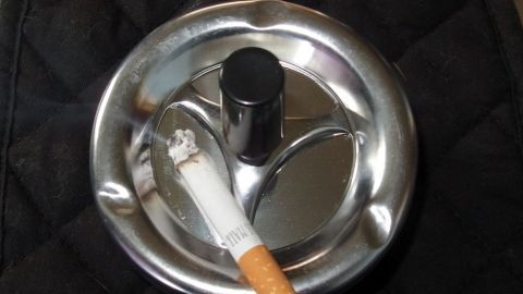 Klamotten stinken nach Rauch? Den Rauchgeruch kann man mit Essig und einem Trockner entfernen. Ein metallischer Aschenbecher mit einer Zigarette liegt darauf, auf einem dunklen, quilted Hintergrund.