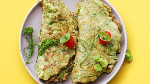 Zucchini-Puffer liegen auf einem grauen Teller, garniert mit Kirschtomaten und frischen Kräutern, auf gelbem Hintergrund.