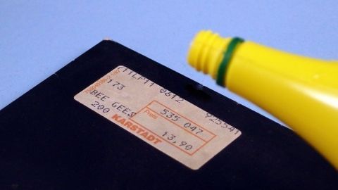 Ein gelbes Flaschenetikett mit Zitronensaft wird über einem Aufkleber auf einem schwarzen Objekt gehalten, um Klebereste zu entfernen.