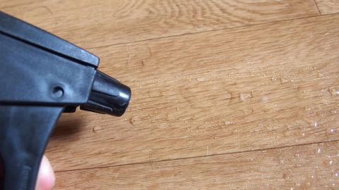 Ein Sprühgerät zielt auf einen hölzernen Boden, der bereit zum Wischen ist, während feiner Staub sichtbar ist.