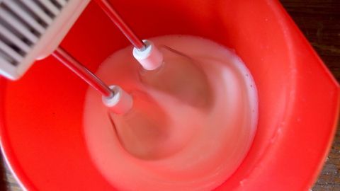 Ein Handmixer rührt cremigen Käsekuchen-Mix in einer roten Schüssel, bereit zum Backen ohne Boden.