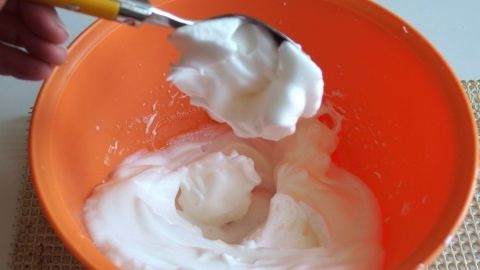 Eine Person hebt mit einem Löffel steifen Eischnee aus einer orangefarbenen Schüssel, um ihn weiterzuverarbeiten.
