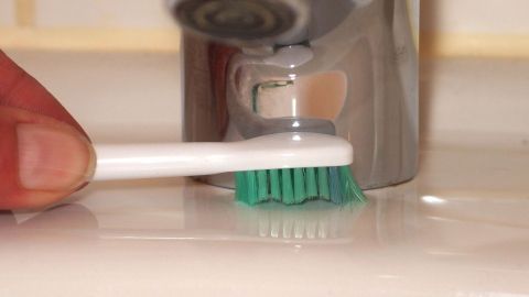 Alte Zahnbürsten eignen sich hervorragend zum Schrubben von kleinen Winkeln und Ecken im Bad. Eine Zahnbürste wird in der Nähe eines Wasserhahns auf einer Waschbeckenablage benutzt, um Zähne zu putzen.