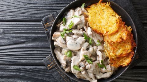 Ein Teller mit Creme-fraiche-Geschnetzeltem und knusprigen Rösti, garniert mit frischem Grün, auf einer dunklen Holzoberfläche.