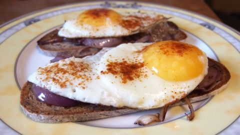 Spiegeleier mit Gemüsezwiebeln liegen auf dunklem Brot, angerichtet auf einem weißen Teller, umgeben von einem rustikalen Tisch.