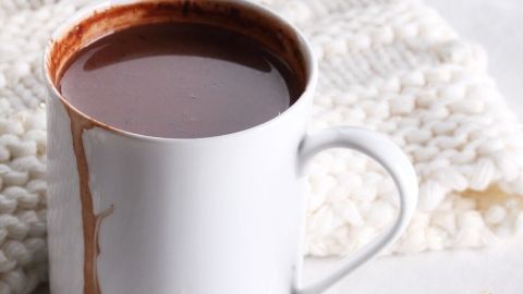 Eine Tasse mit heißem Schokoladengetränk steht auf einem weißen, gestrickten Tuch, daneben liegt ein kleine Löffel.