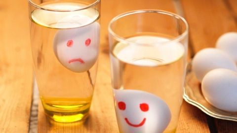 Ein Ei mit einem traurigen Gesicht schwimmt in einem Glas Wasser, während ein fröhliches Ei in einem anderen Glas schwimmt.