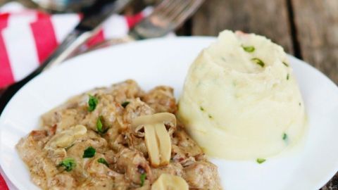 Ein Teller Boeuf Stroganoff liegt neben cremigem Kartoffelbrei auf einem rustikalen Holztisch.