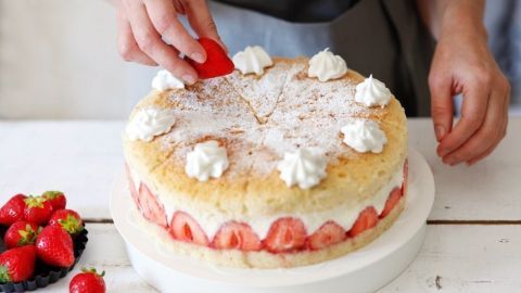 Eine Person dekoriert einen Tortenboden mit frischen Erdbeeren und Sahne, alles auf einem Holztisch angerichtet.