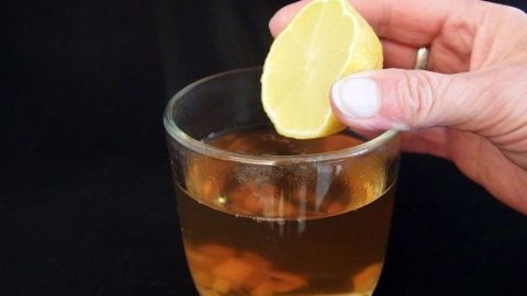 Ein Glas Erkältungstrunk wird zubereitet, während eine Hand eine Zitronenscheibe hineinlegt, auf dunklem Hintergrund.
