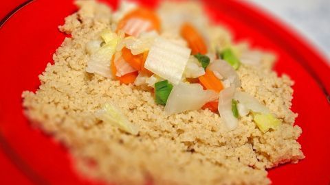 Leckerer Gemüse-Couscous liegt auf einem roten Teller. Bunte, geschnittene Gemüsewürfel sind darüber verteilt.