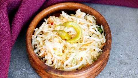Ein Holzschälchen mit frischem, gehobeltem Krautsalat und einer grünen Chili, auf einer grauen Oberfläche serviert.