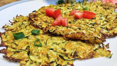 Zucchinipuffer liegen auf einem Teller, garniert mit Tomatenstücken und Frühlingszwiebeln, serviert in einer gemütlichen Küche.