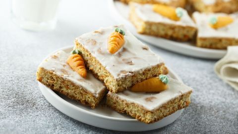 Ein saftiger Möhrenkuchen liegt auf einem Teller, dekoriert mit kleinen Karotten aus Zuckerguss, umgeben von einer hellgrauen Oberfläche.