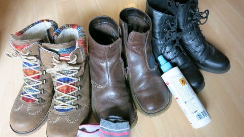Bunte Stiefel und Schuhe stehen auf einem Boden, zusammen mit Socken und einem Pflegeprodukt, um Fuß- und Nagelpilz zu vermeiden.