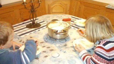 Kinder essen Milchreis an einem dekorierten Tisch in einer Holzbank-Ecke mit Kerzenständer, während ein großer Topf in der Mitte steht.