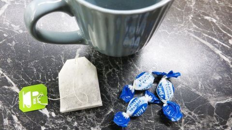 Ein Tee in einer Tasse steht neben einem Teebeutel und Hustenbonbons auf einer dunklen Oberfläche.