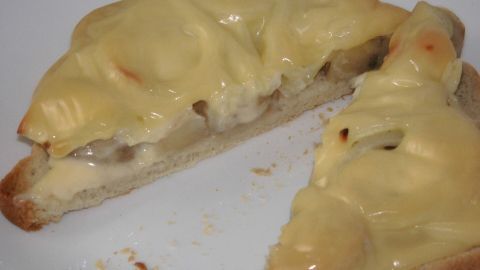 Ein warmes, belegtes Brotstück liegt auf einem Teller, mit geschmolzenem Käse überzogen, serviert in einer einfachen Küche.