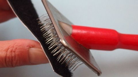 Verfusselte Klettverschlüsse reinigen mit einer Drahtbürste für Tierfellpflege. Ein Handwerkzeug mit rotem Griff reinigt einen Klettverschluss von Fusseln auf einer blauen Oberfläche.