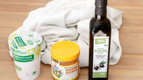 Eine Joghurtpackung, Honig und Olivenöl stehen auf einem Holzuntergrund neben einem Handtuch, bereit für eine Kopfhautbehandlung.