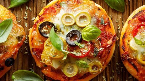 Kleine Pizzabrötchen liegen frisch gebacken auf einem Holzbrett, garniert mit Oliven, Tomaten und frischem Basilikum.