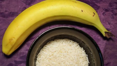 Eine Banane liegt neben einer Schüssel mit gekochtem Reis auf einem lila Hintergrund, ideal bei Durchfall.