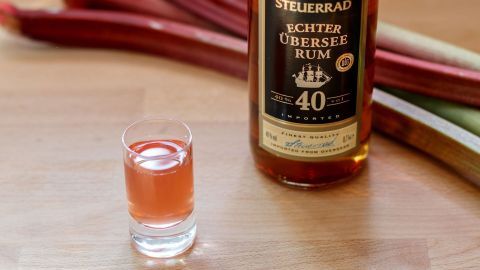 Ein Obstler steht neben einem kleinen Glas, gefüllt mit Rhabarberschnaps, auf einem Holztisch, umgeben von Rhabarberstangen.