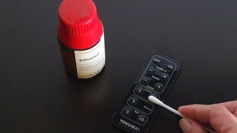 Ein Personenhand greift mit einem Wattestäbchen einen verklebten Taschenrechner, während daneben eine Flasche Reinigungsmittel steht.