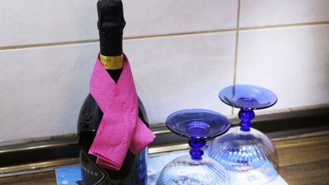 Eine Sektflasche mit einem pinkfarbenen Tuch steht neben zwei blauen Gläsern auf einem Tisch in einer hellen Umgebung.